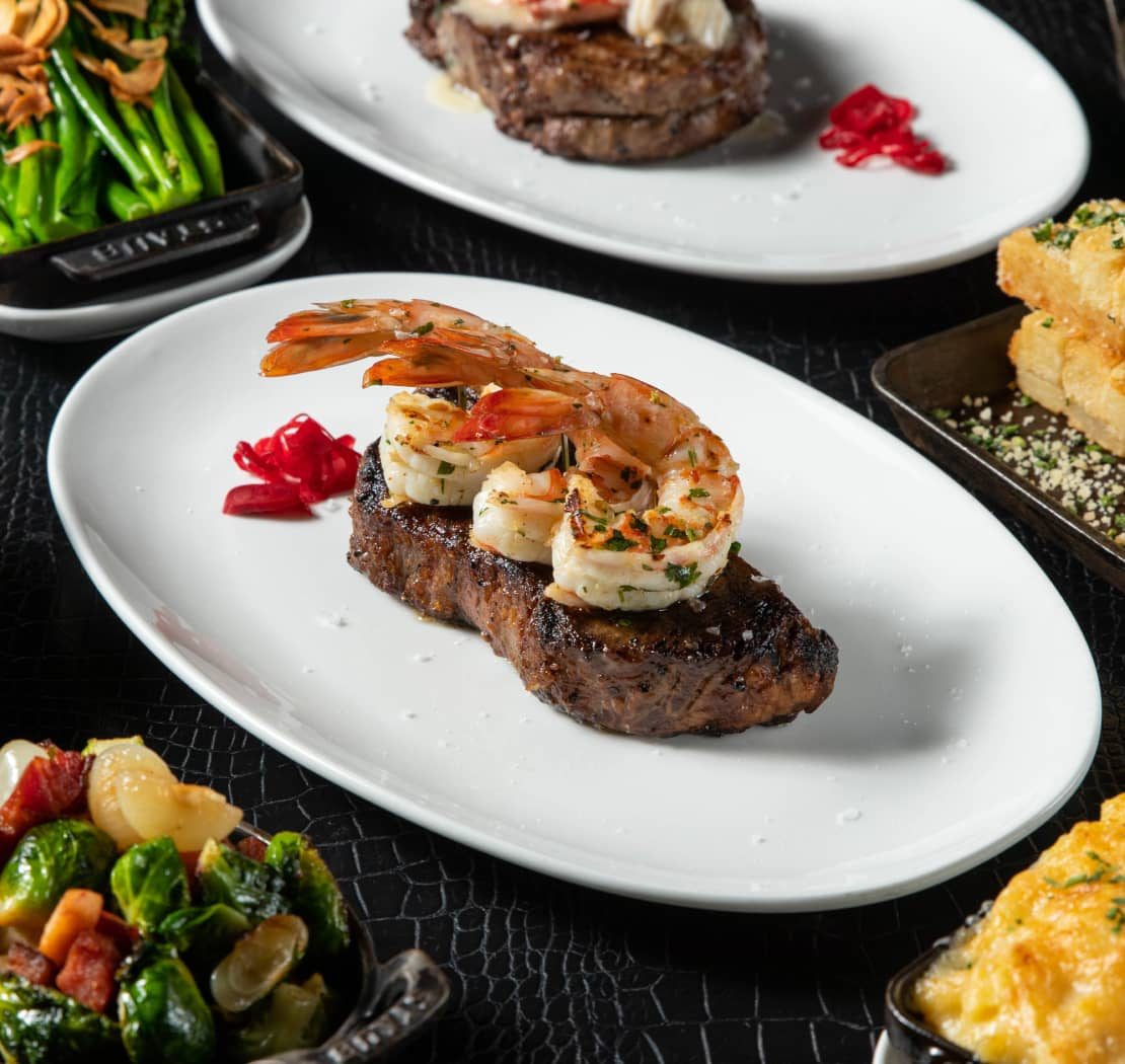 Steak topped with grilled prawns on a white plate, with side dishes