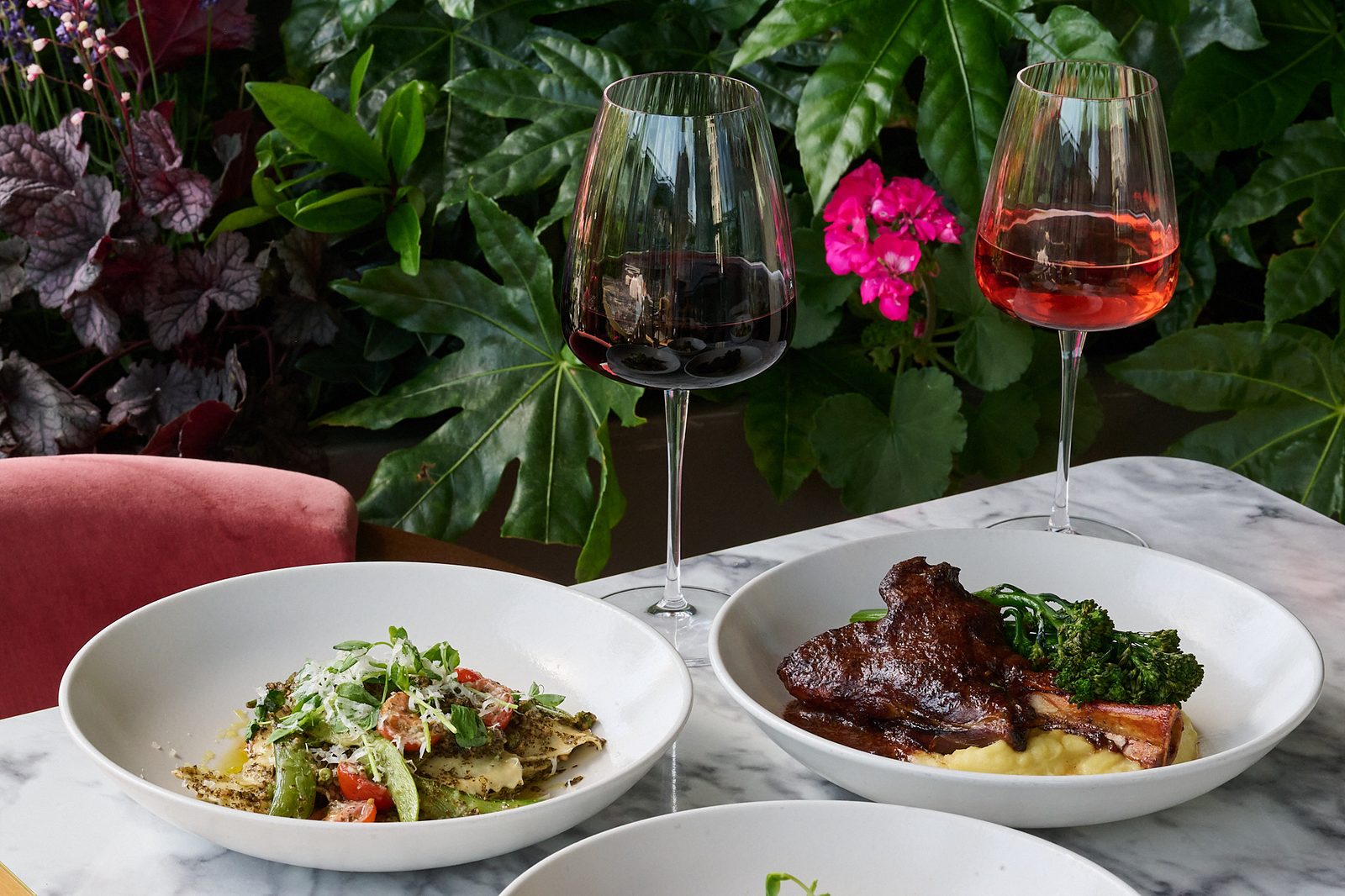 Pasta and a braised meat dish on mash with broccoli, beside glasses of red and rosé wine on a marble table