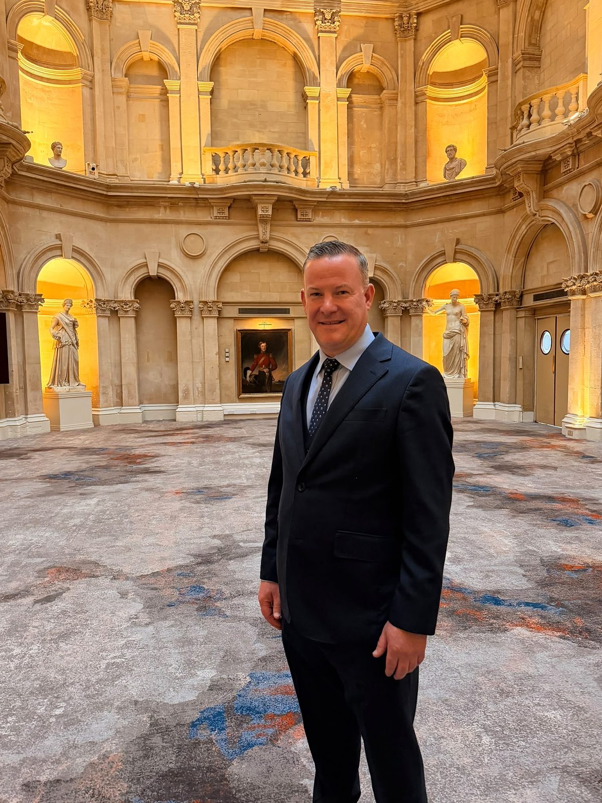 Adam Flint standing in a hall with arched niches, classical statues, a portrait painting and a patterned carpet