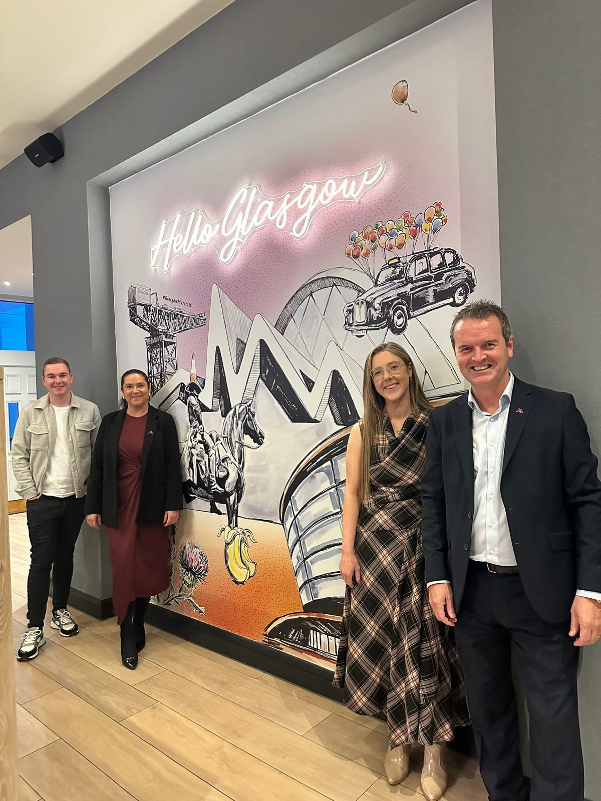 Four members of the Glasgow Marriott Hotel team standing beside the ‘Hello Glasgow’ mural in the hotel lobby