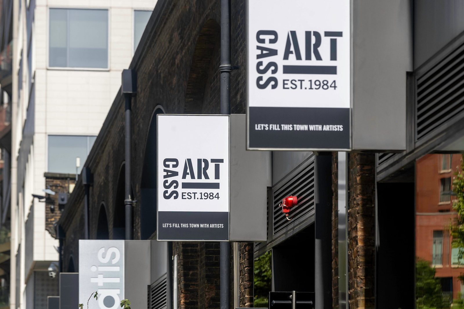 Cass Art projecting signs reading Let’s fill this town with artists, mounted on a building beside brick railway arches