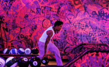 Bowler on a Lane7 lane in front of a pink and purple graffiti wall, with bowling balls on the return
