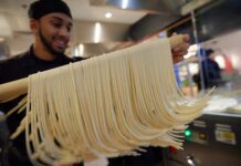 A Marugame Udon team member lifting freshly cut udon noodles on a wooden pole in an open kitchen