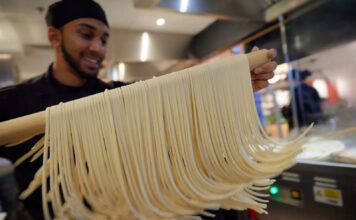 A Marugame Udon team member lifting freshly cut udon noodles on a wooden pole in an open kitchen