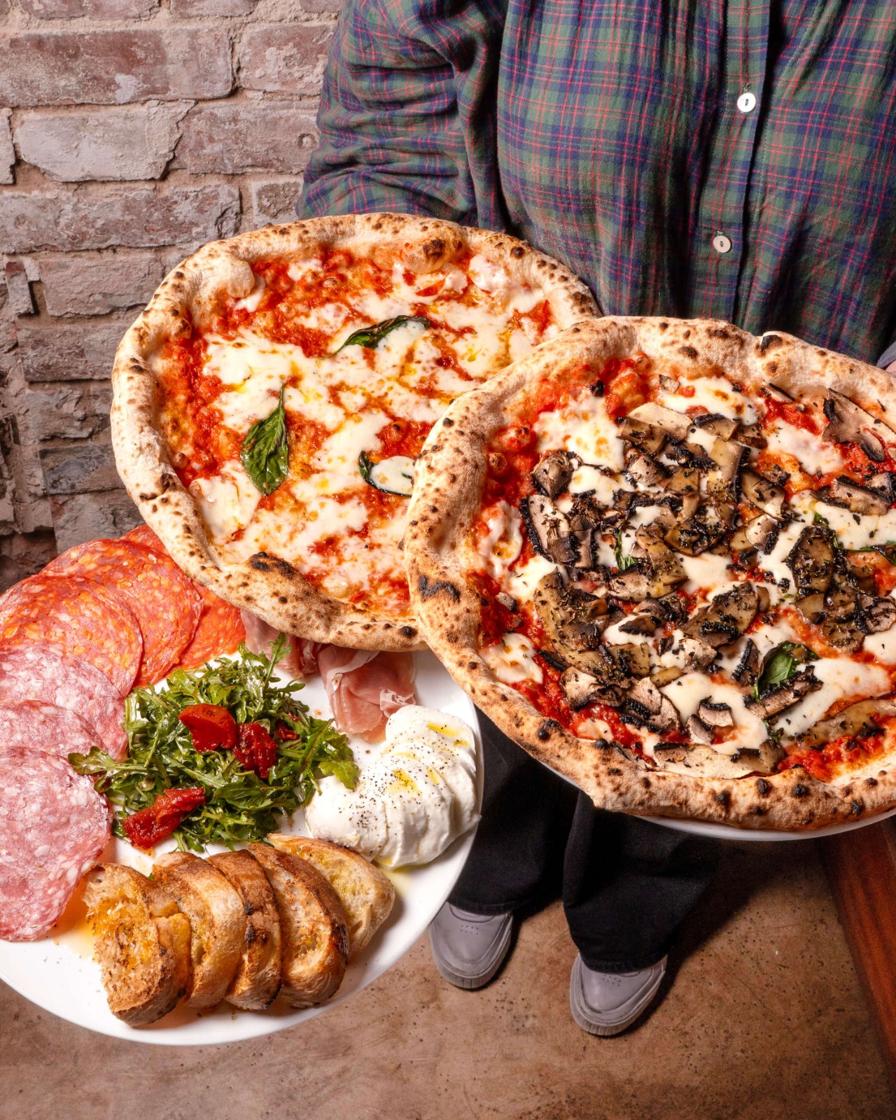 Two Neapolitan pizzas held above a board of cured meats and bread at Rudy’s Pizza Napoletana