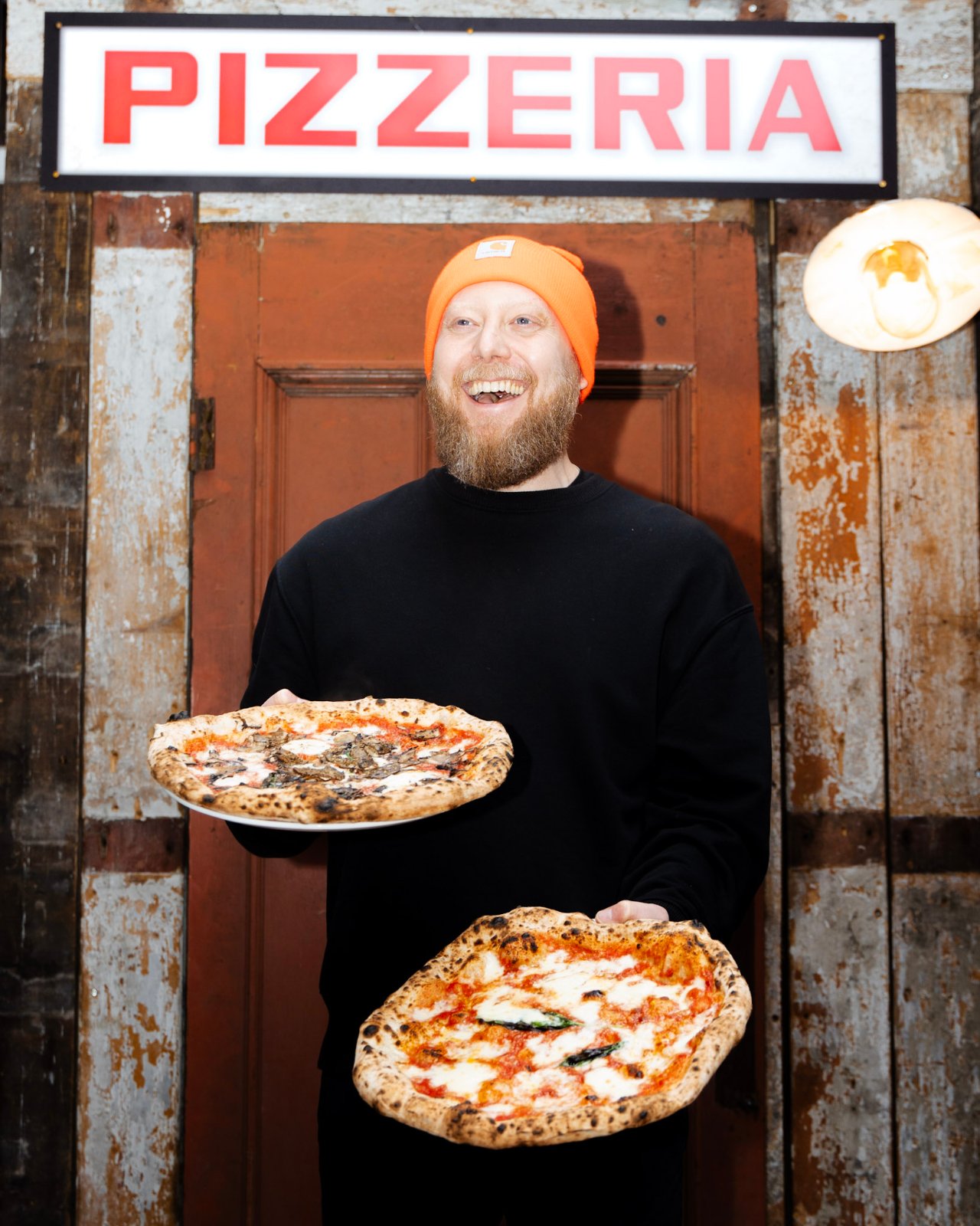 A Rudy’s pizzaiolo holding two pizzas beneath a Pizzeria sign