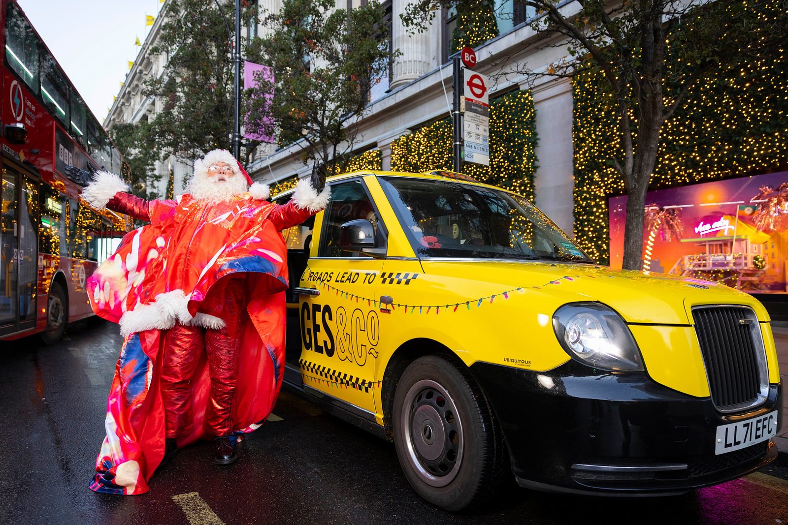 Santa with arms outstretched beside a yellow taxi outside Selfridges on Oxford Street, in front of the Vice City window