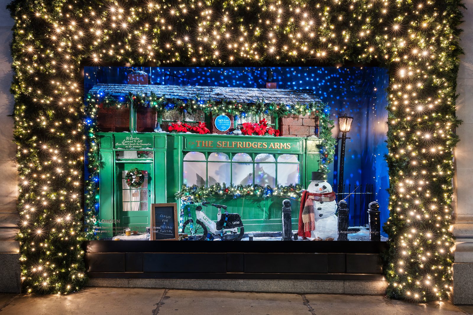 Selfridges' London Christmas window, a green pub front called The Selfridge's Arms with a snowman, framed by a lit garland
