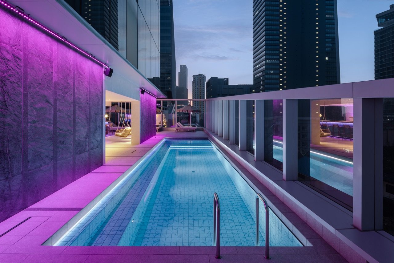 Outdoor pool lit in purple at dusk, with loungers and city towers beyond, at YOTEL Bangkok Sukhumvit